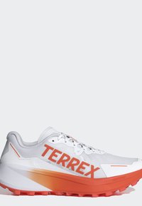 Valkoinen ja oranssi Adidas Terrex juoksukenkä nyörityksellä ja kuvioidulla pohjalla, joka tarjoaa pitoa poluilla, sivulla näkyvä logo.