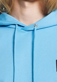 Světle modrá mikina s kapucí vyrobená z měkké bavlněné tkaniny, s tkaničkami se stříbrnými kovovými konci a černým akcentem na lemování.