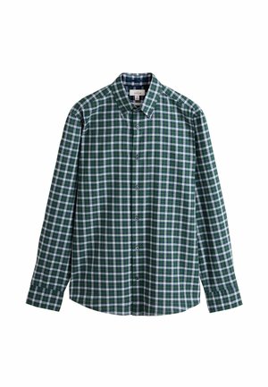 Mann steht drinnen, trägt ein grün-weiß kariertes Hemd, blaue Jeans und braune Loafers mit Quasten, Hand in der Tasche.