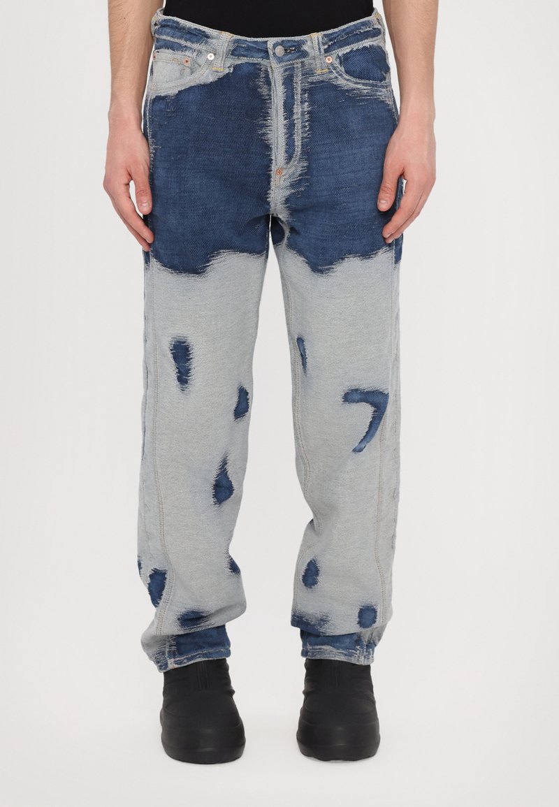 Jean en patchwork bleu et gris avec des motifs usés, coupe décontractée, fermeture à bouton métallique et détails de surpiqûres contrastées.