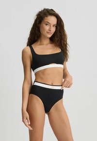 Crni bikini gornji dio s širokim trakama i bijelom trakom; visoki donji dio s kontrastnom bijelom trakom i zlatnim logotipom. Glatka tekstura.