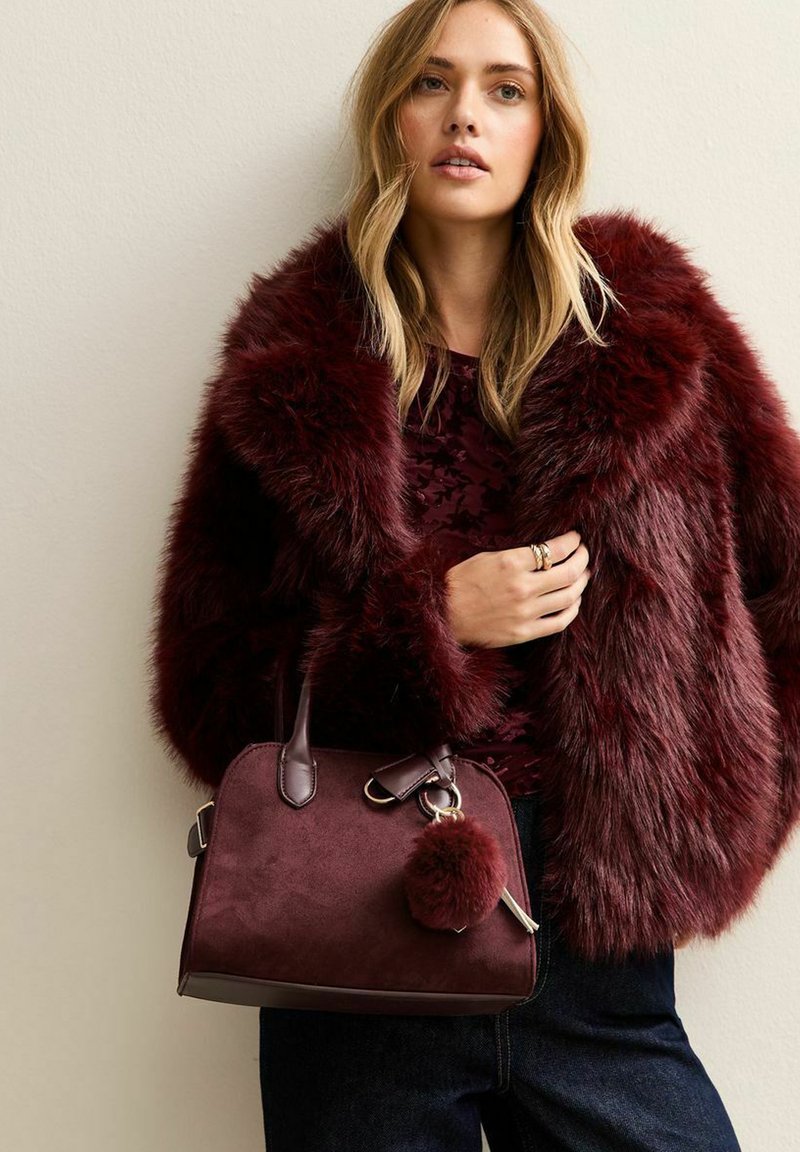 Giacca in pelliccia finta borgogna sopra un top a fantasia, con una borsa bordeaux dotata di un portachiavi con pompon e design strutturato.