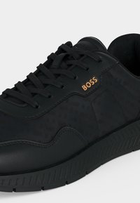 Baskets en cuir et tissu noir avec lacets noirs, semelle texturée et logo doré "BOSS" sur le côté.