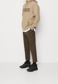 Beżowa bluza z kapturem z przednią kieszenią i czarnym napisem, zestawiona z brązowymi spodniami z mankietami oraz czarnymi trampkami o teksturowanej powierzchni.