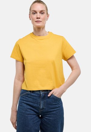 Femme à la peau claire et aux cheveux blonds, portant un t-shirt court jaune à manches courtes et un jean taille haute bleu, debout avec une main dans la poche.