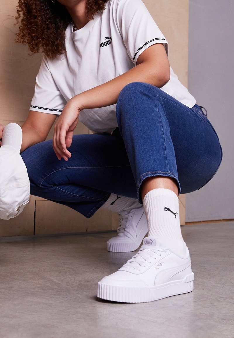 Vita PUMA-sneakers med en tjock, räfflad sula, matchade med vita strumpor som har en svart logotyp. Modellen bär en vit T-shirt och blå jeans.