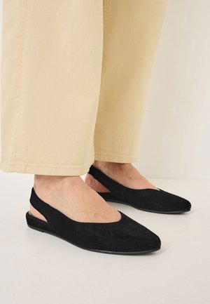 Mustat mokkapintaiset slingback-kengät, joissa on terävä pää, sivuaukot ja litteä pohja, yhdistettynä vaaleisiin, väljiin housuihin.