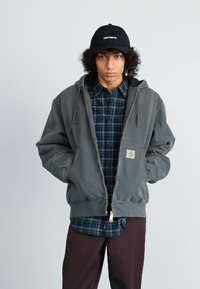 Siva zip-up hoodie s kapuljačom i prednjim džepovima, složena preko plave karirane košulje, nosi se uz smeđe hlače i crnu kapu.