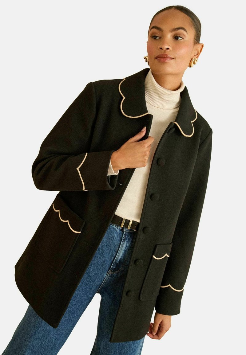 Zwarte wollen mantel met scalloped kraag en crèmekleurige bies, voorzien van twee voorzakken en knoopsluitingen. Gedragen over een lichte coltrui.