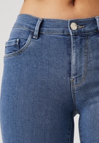Jeans de mezclilla azul con corte ajustado, que cuentan con un cierre estándar de cremallera y botón, bolsillos frontales y detalles de costura visibles.