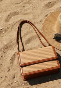 Πλεγμένη τσάντα με καφέ δερμάτινη διακόσμηση και λουριά στην αμμώδη παραλία δίπλα σε ψάθινο καπέλο με μεγάλη γείσο και σκούρα γυαλιά ηλίου.