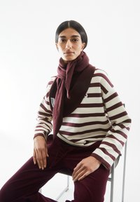 Bordeaux sjaal om de hals, gestreept bordeaux en witte lange mouw shirt, corduroy bordeaux broek, zittend op een metalen stoel.