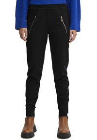 Črne trenirke z elastičnim pasom, dvema zadrgo zaprtima sprednjima žepoma, oprijet fit in gladko teksturo tkanine. V družbi rjavih gležnjarjev.