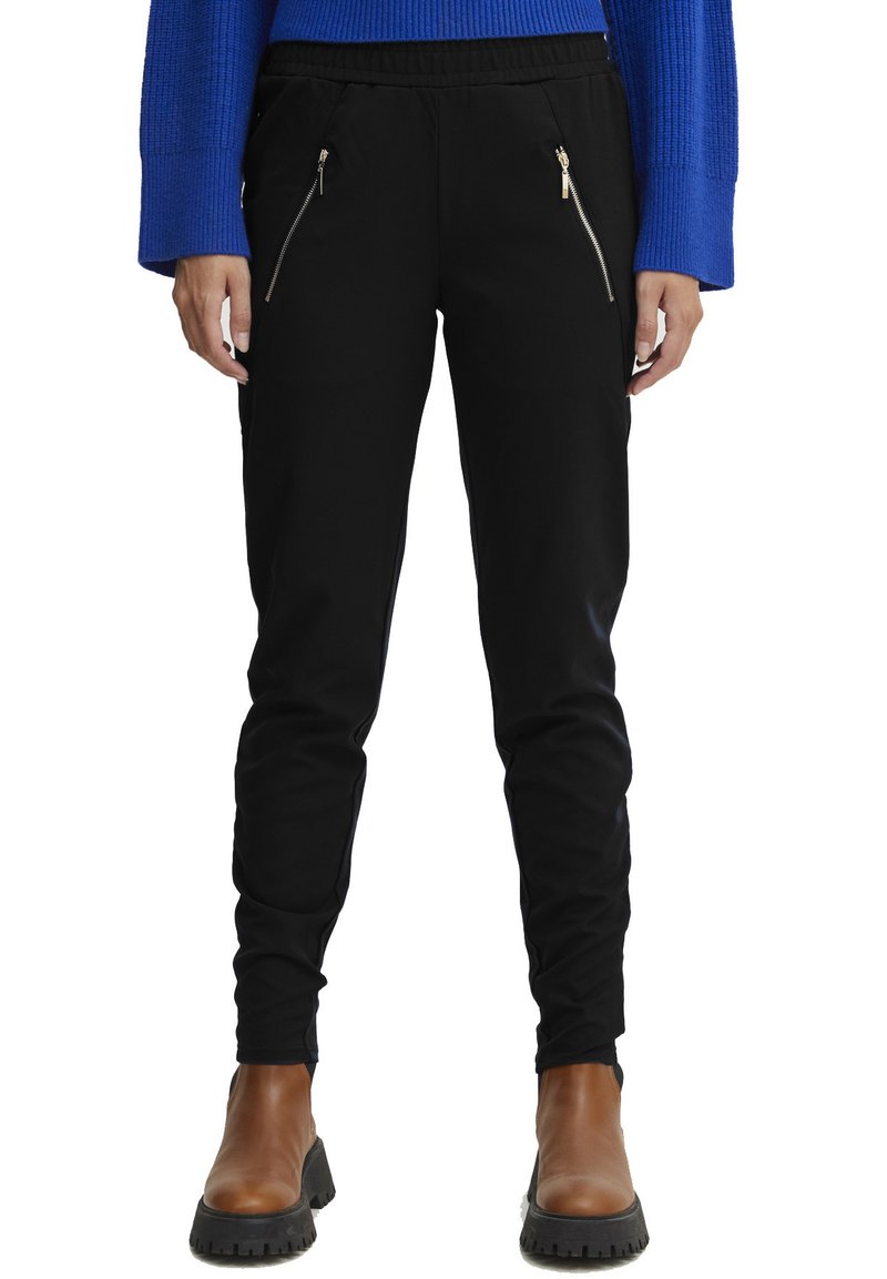 Črne trenirke z elastičnim pasom, dvema zadrgo zaprtima sprednjima žepoma, oprijet fit in gladko teksturo tkanine. V družbi rjavih gležnjarjev.