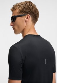 Black short-sleeve athletic shirt with a textured design, featuring a small logo on the back and paired with dark sunglasses.