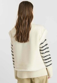 Crème-kleurige jas met een fuzzy textuur, voorzien van gestreepte zwart-witte mouwen en een geribde zoom. De achteraanblik toont een ontspannen pasvorm.