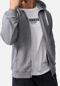 Veste à fermeture éclair grise avec une poche kangourou, une capuche réglable avec cordon de serrage et des poignets côtelés. Portée sur un t-shirt blanc à motif.