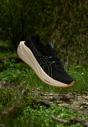 Chaussure de course noire avec une semelle blanche flottant au-dessus d’un chemin de terre dans une forêt verte, montrant le logo de la marque et les détails texturés de la semelle.