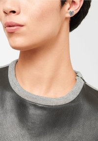 Hjärformade silverörhängen med geometrisk design, framför en modell som bär en svart läderskjorta med en grå krage.