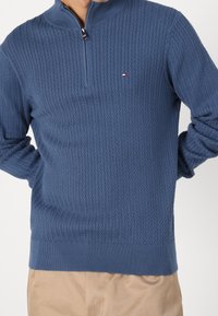 Sininen neulepusero, jossa on vetskari-kaulus ja teksturoitu herringbone-kuvio. Ribatuutiset hihat ja helma, joissa on pieni logo rinnassa.