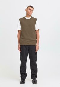 Chaleco de punto verde oliva sobre una camiseta blanca, combinado con pantalones cargo negros. Unos zapatos negros completan el moderno conjunto en capas.