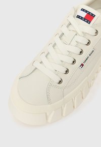 Bijele kožne tenisice s debelim gumenom potplatom, okruglim prstom i istaknutim vezanjem. Imaju branded oznaku i detalje šivanja sa strane.