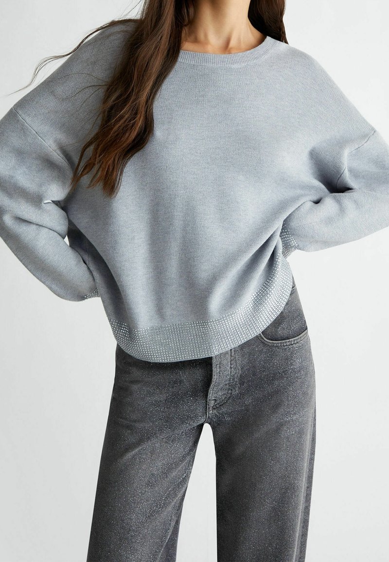 Femme portant un pull à manches longues gris clair avec un ourlet orné et un jean noir délavé, debout les mains sur les hanches devant un fond blanc.