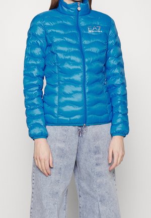 Personne portant une doudoune bleu vif EA7 Emporio Armani et un jean large délavé, debout devant un fond uni.