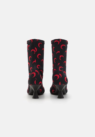 Botas negras hasta la rodilla con patrones de luna creciente roja, hechas de tela elástica. Presentan un tacón negro delgado y angular para mayor estabilidad.