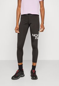 Černé legíny s bílým logem "THE NORTH FACE" na levém stehně, spárované se světle fialovým zkráceným tričkem a černou obuví.