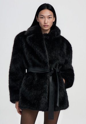 Donna con lunghi capelli neri che indossa un cappotto di pelliccia ecologica nera con un ampio colletto e una cintura nera annodata, in piedi davanti a uno sfondo chiaro e uniforme.