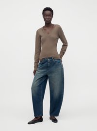 Femme debout portant un pull marron à col en V, un jean bleu ample et des ballerines noires sur un fond blanc uni.