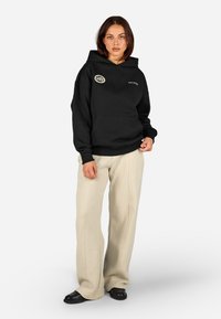 Μαύρη κουκούλα με μπροστινή τσέπη και λογότυπο, συνδυασμένη με υφασμάτινο μπεζ παντελόνι και μαύρα παπούτσια, σε ένα ουδέτερο φόντο.