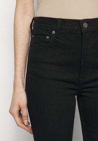 Vänster hand vilande vid sidan av svarta högmidjade jeans, som bärs med en beige ribbad topp, vilket visar jeansens knapp, fickor och bältesöglor.