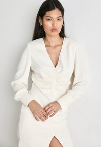 Cremefarbenes Kleid mit V-Ausschnitt, langen Ärmeln, Raffung an der Taille und einem schulterlangen Schlitz. Glattes Material mit subtiler Textur.