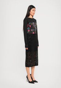 Black long-sleeve top with floral embroidery and a fitted design, paired with a long black lace skirt. Black high-heeled shoes complete the look.