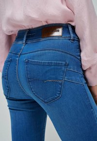 Jeans de mezclilla azul con un diseño ajustado, que presentan bolsillos traseros y una etiqueta de marca en bronce. Detalles de bordado sutil a lo largo de los bordes de los bolsillos.