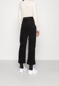 Pantalon large en velours côtelé noir avec poches arrière, porté avec un haut à manches longues crème et des baskets blanches. Texture visible sur le tissu.