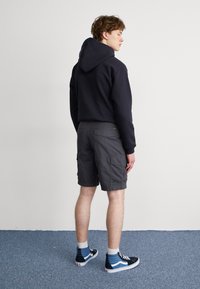 Sudadera negra con capucha combinada con pantalones cortos de carga grises. El modelo lleva zapatillas altas en azul y negro, mostrando paneles y texturas distintivas.