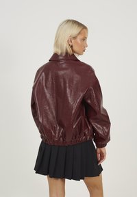 Giacca in ecopelle bordeaux con vestibilità corta e ampia, vita elasticizzata e colletto a punta; il modello indossa una gonna nera plissettata.