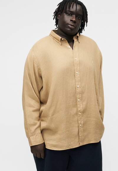 Vyro su tamsia oda ir pintais plaukais, dėvinčio laisvą smėlio spalvos mygtukais užsegamą marškinių viršų ir tamsias kelnes, stovinčio priešais paprastą šviesią foną.