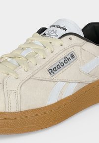 Μπεζ σουέτ αθλητικό παπούτσι Reebok με λευκές ρίγες, κρεμ κορδόνια, σόλα από καουτσούκ σε χρώμα τσίχλας και μαύρη εσωτερική επένδυση, εμφανίζεται από το πλάι σε κοντινή λήψη.