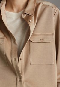 Chemise beige à boutons en tissu doux, avec un col pointu, deux poches poitrine et un léger éclat. Pull à col rond en dessous.