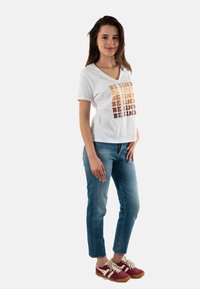 Camiseta de algodón blanca con escote en V, con la frase "SE AMABLE" impresa en tonos marrones degradados. Lleva puesta unos vaqueros azules y zapatillas burdeos.