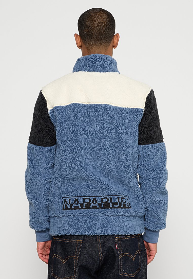 Hombre de espaldas que lleva una chaqueta de forro polar azul, blanca y negra con el texto "NAPAPIJRI" en la parte baja de la espalda y vaqueros de mezclilla oscuros.