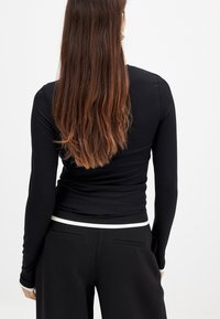 Svart långärmad topp i slät tyg med en skräddarsydd design. Har ett vitt accentband vid midjan. Bakifrån syns hår som faller ner.