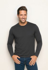 Langärmliges schwarzes Baumwollhemd mit Rundhalsausschnitt, glatter Textur und dezenten Nähten, kombiniert mit blauen Jeans und einem neutralen Hintergrund.