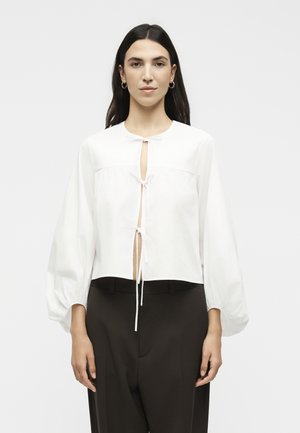 Γυναίκα με μακριά σκουρόχρωμα μαλλιά φορώντας λευκό πουκάμισο με μακριά μανίκια δεμένο μπροστά και σκουρόχρωμο παντελόνι, κοιτάζει μπροστά σε απλό φόντο.