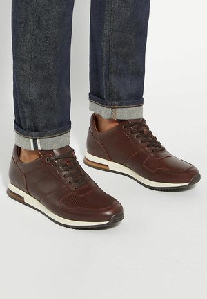 Brown leather sneakers with textured panels, flat laces, rubber soles, and cream accents. Jeans with rolled cuffs complement the footwear.