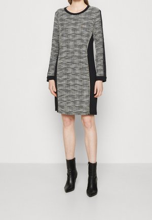 Femme portant une robe à motifs noire et grise mi-longue à manches longues, avec des bottines noires, debout devant un fond uni.